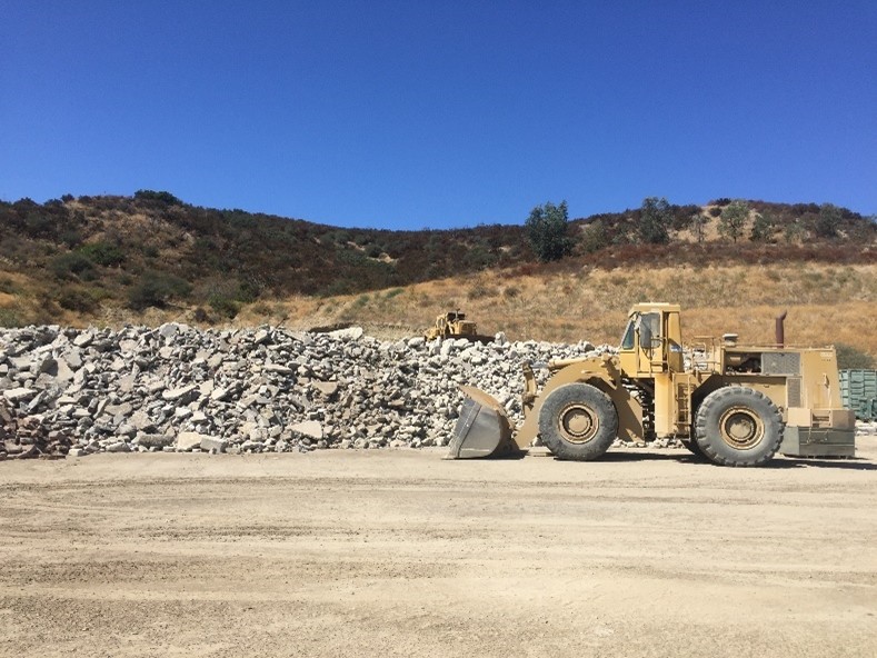 Large bulldozer beside a pile of rocks at a quarry site, with hills in the background.